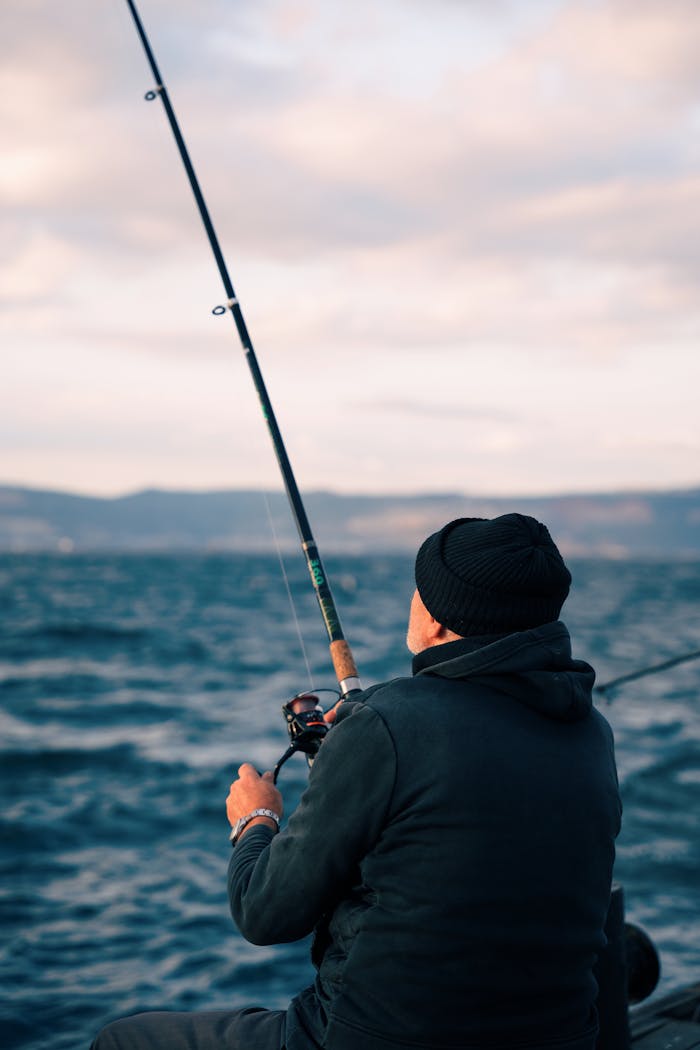 Back view of a man fishing at sea, casting a line with a serene ocean and sky backdrop.
