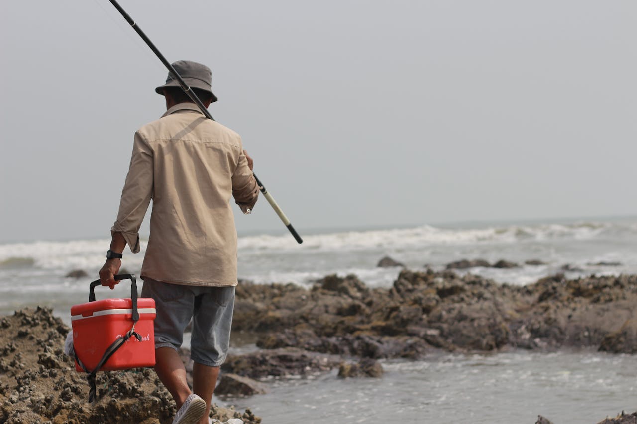 A lone fisherman walks along the rocky shore carrying fishing gear and a cooler.