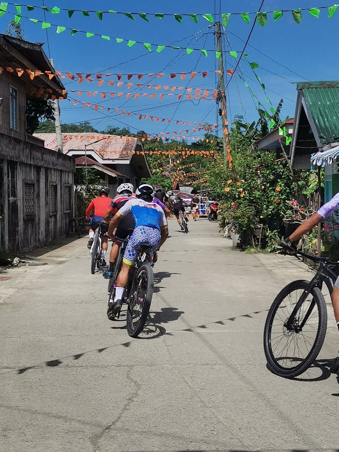 creative-03 Cyclists ride through a village street adorned with colorful flags under a clear blue sky.