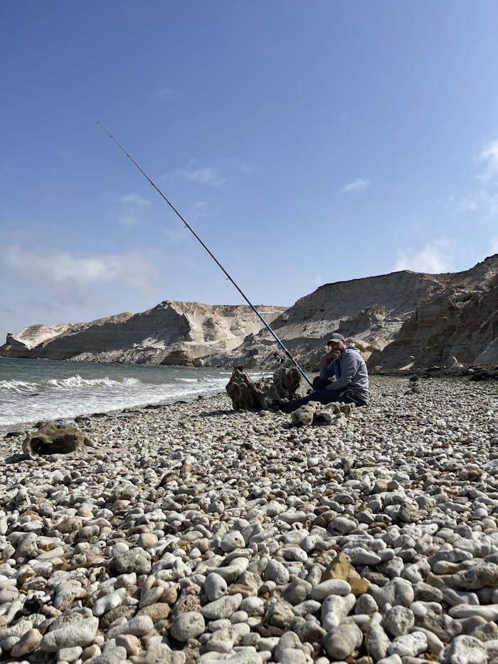 A lone fisherman sits on a rocky beach casting a line into the ocean under a clear blue sky.